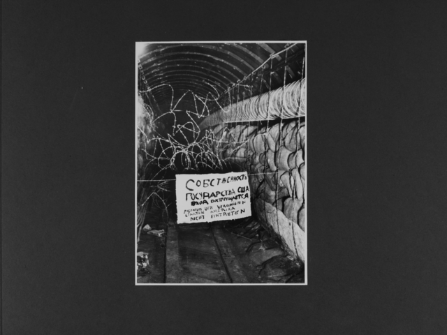[Das Lichtbild zeigt einen langen Tunnelgang, der auf beiden Seiten gestapelte Jutesäcke hat. Der Eingang ist durch Stacheldraht auf ganzer Höhe versperrt. Davon hängt ein Schild, auf dem in schwarzer Farbe zuerst mit kyrillischen Buchstaben sowie darunter in Deutscher Sprache "Eigentum der Vereinigten Staaten Amerika; nicht eintreten" geschrieben steht.]