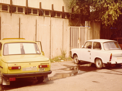 Auf einer betonierten Fläche stehen ein hellgrüner Wartburg und, mit einer Parklücke dazwischen, ein papyrusfarbener Trabi. Sie stehen vor einer Betonmauer und mit dem Heck zum Fotografierenden. Es ist ein farbiges Lichtbild.