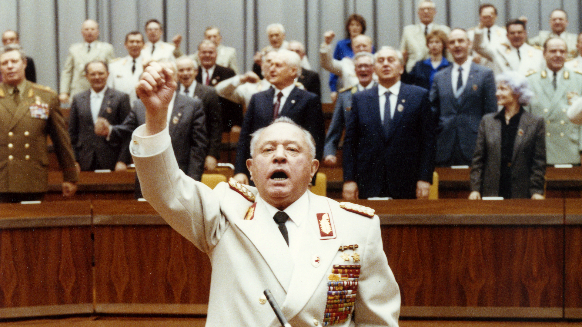 Das Bild zeigt Erich Mielke an einem Mikrofon mit erhobener rechter Faust.