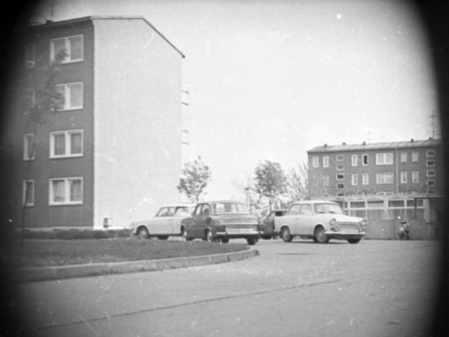 Zwischen zwei viergeschossigen Neubaublöcken befindet sich ein Flachbau mit großen Fenstern, bei dem es sich vermutlich um eine gastronomische Einrichtung handelt. Der Schriftzug auf dem Dach ist nicht zu erkennen. Im Vordergrund parken mehrere Pkw und ein Moped.
