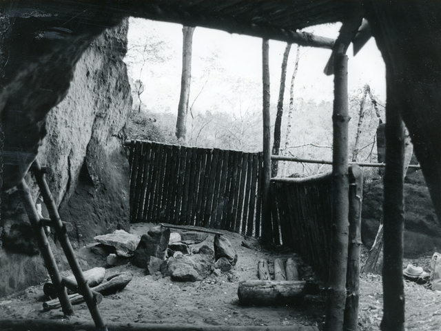Hierbei handelt es sich um eine Schwarzweißfotografie des Ausblickes aus dem Inneren der Boofe. Man blickt nach vorne und zur rechten auf einen hüfthohen Zaun, hinter dem Baumwipfel zu erkennen sind, links befindet sich die Felswand. Auf dem Boden ist eine Feuerstelle aufgebaut, neben der Holzscheite und Felsbrocken lagern, während links eine kleine Leiter am Gestein angelehnt steht. 