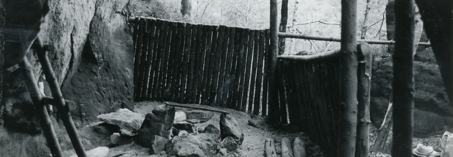 Hierbei handelt es sich um eine Schwarzweißfotografie des Ausblickes aus dem Inneren der Boofe. Man blickt nach vorne und zur rechten auf einen hüfthohen Zaun, hinter dem Baumwipfel zu erkennen sind, links befindet sich die Felswand. Auf dem Boden ist eine Feuerstelle aufgebaut, neben der Holzscheite und Felsbrocken lagern, während links eine kleine Leiter am Gestein angelehnt steht. 
