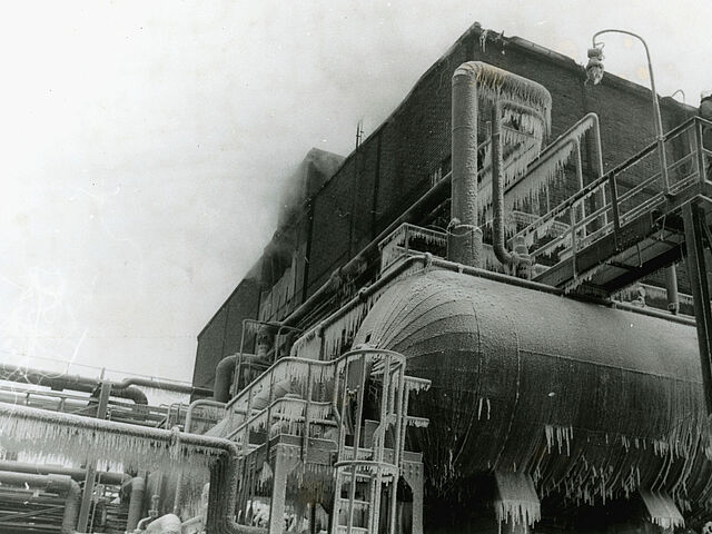 [Auf der schwarz-weiß Fotografie ist ein Teil des Industriegebäudes von Bild 17 zu sehen, aus dessen Stirnseite Dampf emporsteigt. Links neben dem Gebäude verlaufen einige Rohre. Vor dem Gebäude ist ein waagerechter Tank mit Leiter zu sehen. Über dem Tank führt eine Treppe hinunter. Auf dem obersten Absatz der Treppe ist ein Mann mit Schutzhelm zu erkennen. Da die Rohre und der Tank mit vielen Eiszapfen überzogen sind, wurde das Foto vermutlich im Winter aufgenommen.]