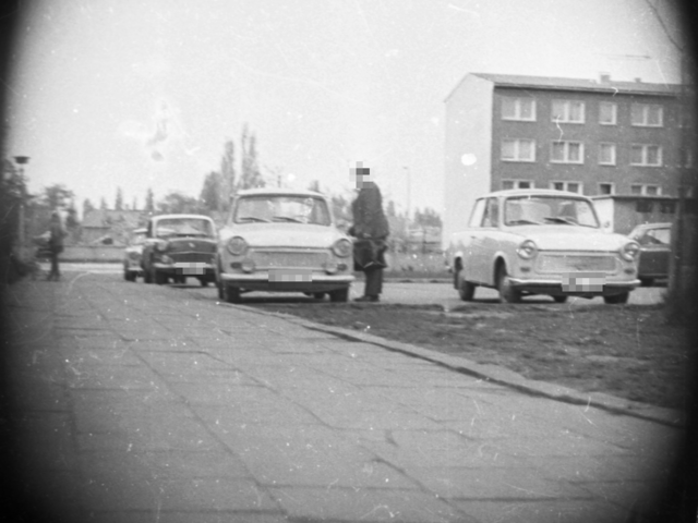 Mit versteckter Kamera wurde ein Mann fotografiert, der eine Tasche in der Hand hält und an der Fahrertür eines Trabants steht. Rundherum stehen weitere Pkw. Der Parkplatz befindet sich neben einem viergeschossigen Mehrfamilienhaus.