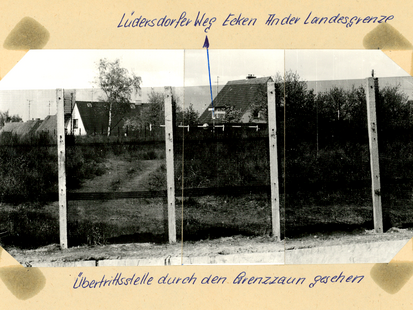[Hinter dem Grenzzaun wurde der Grünstreifen mit angrenzenden Einfamilienhäusern fotografiert. Ein unbefestigter Weg führt in Richtung der Häuser (dort an einer Schranke endend) sowie an den Grenzzaun. Es handelt sich um ein schwarzweißes Lichtbild, auf dem mit blauem Kugelschreiber per Pfeil eine Markierung vorgenommen wurde. Die Beschriftung folgt außerhalb des aufgeklebten Fotos auf Papierblatt:] Lüdersdorfer Weg Ecken An der Landesgrenze  [handschriftlich mit blauem Kugelschreiber: Übertrittsstelle durch den Grenzzaun gesehen]