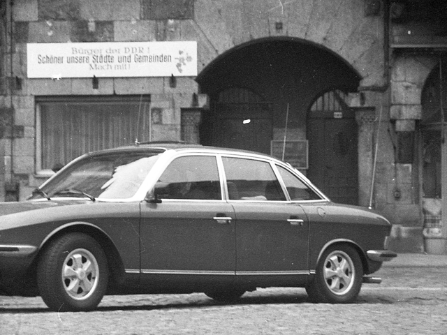 [Auf einem gepflasterten Platz steht ein Pkw, vermutlich ein NSU Ro80. Im Hintergrund ist ein Stadthaus mit schmuckreicher Fassade zu sehen. Neben einer doppelten Rundbogentür hängt ein schmales Schild über einem Fenster mit der Aufschrift "Bürger der DDR! Schöner unsere Städte und Gemeinden Mach mit!" Vermutlich wurde die Aufnahme verdeckt getätigt da der Bildausschnitt von einem runden dunkleren Rahmen umgeben sind. Das Original ist ein schwarz-weißes Negativ.]