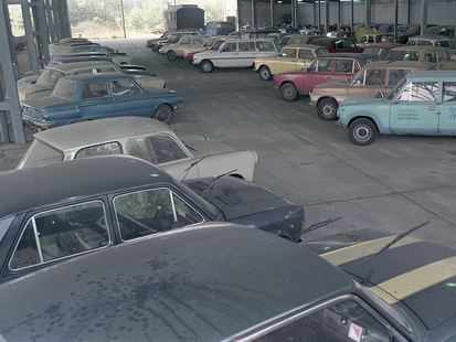 Ein überdachter Betonplatz wurde als Parkplatz für verschiedenfarbige Modelle von Pkws benutzt. Die Autos stehen in Schrägaufstellung und es gibt nur wenige Lücken. Alle Scheibenwischer wurden von den Frontscheiben weggeklappt. Auf der linken Bildseite sind ein paar Stützpfeiler sowie ein Maschendraht zu sehen, doch auch dahinter stehen Kraftfahrzeuge. Es ist ein weitwinklig aufgenommenes, farbiges Lichtbild und wurde von leicht erhöhter Position aufgenommen.