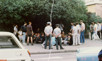 Eine Personengruppe sitzt auf einem Bürgersteig direkt vor einem Zaun. Sie werden umringt von Volkspolizisten sowie einigen steif wirkenden Herren in ordentlichem Zivil. Das farbige Lichtbild wurde manuell rekonstruiert, in der Mitte ist der vertikale Riss sichtbar.]