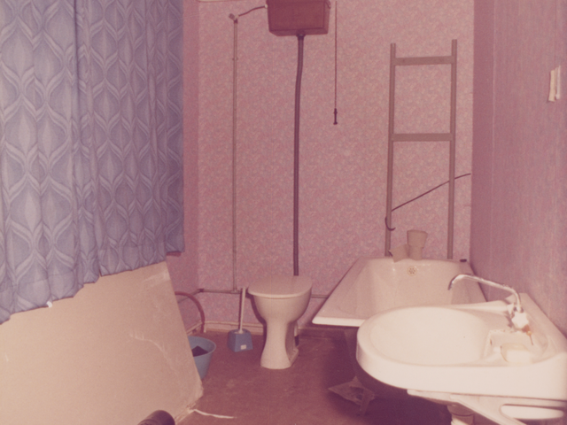 [Auf dem Farbfoto ist ein Badezimmer abgebildet, das mit braunem Linoleum ausgelegt ist. An der rechten Wand hängt vorn ein Waschbecken, dahinter steht eine Badewanne. Links neben der Badewanne steht das WC-Becken, das durch ein Rohr mit dem darüber hängenden Spülkasten verbunden ist. Unter dem Fenster, das sich in der linken Zimmerwand befindet, ist eine Platte an die Wand gelehnt. Vor ihr steht ein röhrenförmiger elektronischer Bahnheizkörper.]