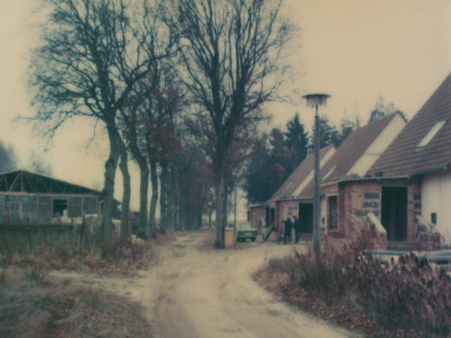 [Die Farbaufnahme zeigt einen Sandweg. Links vom Weg ist eine große Industriehalle zu sehen. Auf halber Bildhöhe zweigt der Sandweg nach rechts ab und führt zu drei nebeneinander angeordneten baugleichen Häusern, deren Wände und Mauern noch unverkleidet und unverputzt sind. Die Spitzdächer sind rot geziegelt. Im Inneren der Wegbiegung steht eine für die DDR typische "Rostocker Straßenleuchte" (RSL 1), daneben liegen aufgestapelt Kanalrohre. Vor dem mittleren Haus parkt ein grüner Trabant. Zwei Männer stehen daneben und unterhalten sich.]