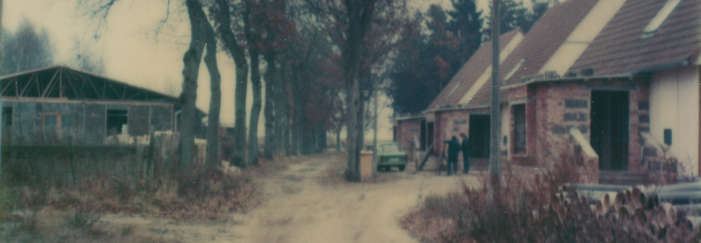 [Die Farbaufnahme zeigt einen Sandweg. Links vom Weg ist eine große Industriehalle zu sehen. Auf halber Bildhöhe zweigt der Sandweg nach rechts ab und führt zu drei nebeneinander angeordneten baugleichen Häusern, deren Wände und Mauern noch unverkleidet und unverputzt sind. Die Spitzdächer sind rot geziegelt. Im Inneren der Wegbiegung steht eine für die DDR typische "Rostocker Straßenleuchte" (RSL 1), daneben liegen aufgestapelt Kanalrohre. Vor dem mittleren Haus parkt ein grüner Trabant. Zwei Männer stehen daneben und unterhalten sich.]
