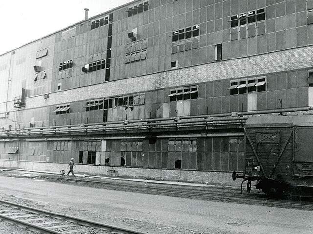 [Auf der schwarz-weiß Fotografie ist ein mehrstöckiges Industriegebäude zu erkennen. Das Gebäude besteht aus vielen Fenstern, einige stehen offen und einige sind beschädigt. Über dem ersten Stock verlaufen einige Rohre parallel. Davor ist ein Mann mit einem Schutzhelm zu sehen. Er läuft in Richtung eines Güterwagons, der rechts vor dem Gebäude steht. Vor dem Gebäude verlaufen zwei Eisenbahnschienen.]