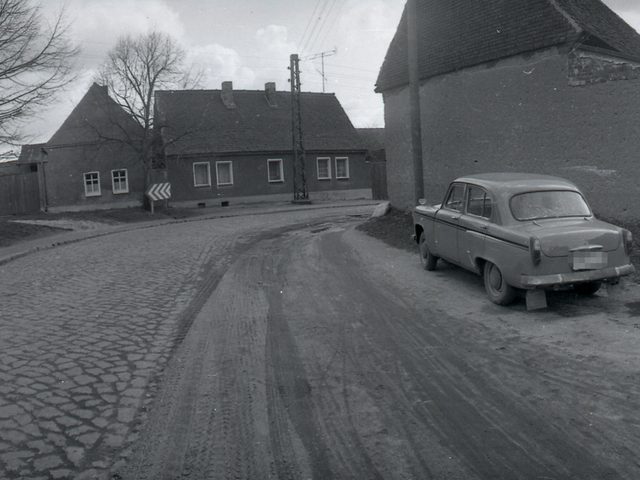 Zu sehen sind eine Reihe von Einfamilienhäusern sowie parkende Autos am Rand einer kopfsteingepflasterten Straße in einer vermutlich ländlich gelegenen Ortschaft.