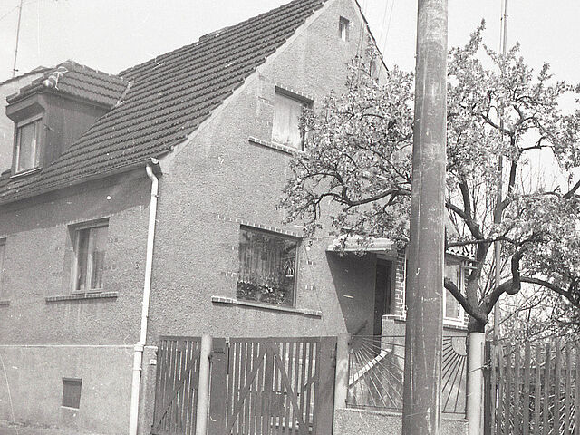 [Die schwarz-weiß Fotografie wurde von der Beifahrerseite eines parkenden Fahrzeuges geschossen. Links auf der Fotografie ist ein Einfamilienhaus an einer Straße zu sehen. Auf dem Grundstück, rechts neben dem Haus, steht ein Kirschbaum. Angrenzend an den Kirschbaum beginnt ein neues Grundstück.]