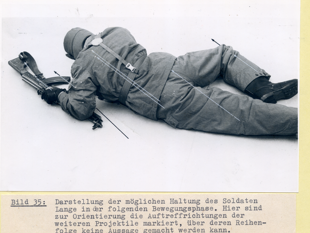 [Auf den beiden schwarzweißen Fotos liegt eine Person, sich mit den Armen hoch drückend, leicht seitlich auf dem Bauch und dreht dabei den Rücken zur Kamera. Dabei ist sie in Uniform und Koppel der NVA-Grenztruppen gekleidet und trägt auch die Fellmütze. Das rechte Bein ist angewinkelt, mit dem linken Arm stützt sie sich auf der Maschinenpistole auf. Dabei ist das Lauf unter dem Unterarm, die restliche Hälfte der Waffe liegt in Richtung des Kopfes und damit von der Kameraposition weg. Es sind feine schwarze und graue Pfeile zu sehen, die die Auftreffrichtungen der Projektile beim Soldaten Lange markieren.]
