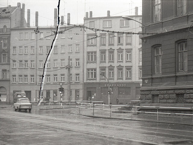 [Eine schwarzweiße Aufnahme einer T-Kreuzung mit Mittelinsel und Ampelsteuerung. Zwei Fahrzeuge sind am linken Bildrand, der Straßenbelag glänzt und war vermutlich nass geregnet. Die runden Straßenecken sind mit Fußgängersperrgittern versehen. Die vermutlichen Wohnhäuser einer Innenstadt sind Altbauten. Das Negativ ist durch einen vertikalen Riss auf der linken Seite beschädigt.]