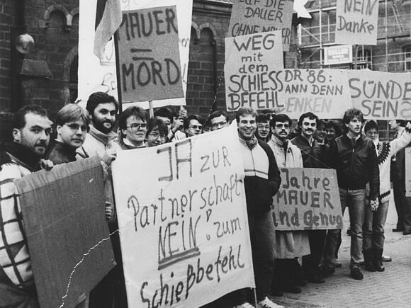 Auf diesem Schwarz-Weiß Foto ist eine Gruppe von Demonstranten zu sehen. Sie halten viele verschiedene Demonstrantionsschilder in den Händen mit Sprüchen wie zum Beispiel "Mauer = Mord", "Ja zur Partnerschaft. Nein zum Schießbefehl" oder "Weg mit dem Schießbefehl" hoch.