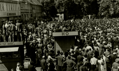 Der Marktplatz des thüringischen Sömmerda am 17. Juni 1953: Im Hintergrund die Menschenmenge, die anscheinend einer Kundgebung beiwohnt. Im Vordergrund die Bretterbuden 'Zeitungsvertrieb' und 'HO, auf denen Menschen sitzen oder stehen. Ganz vorne weitere Menschen, manche von ihnen mit Fahrrädern. Auf der Rückseite des Fotos ist handschriftlich vermerkt: 'Sömmerda am 17.6.53 – Demonstranten auf dem Marktplatz in Sömmerda (Rheinmetall).