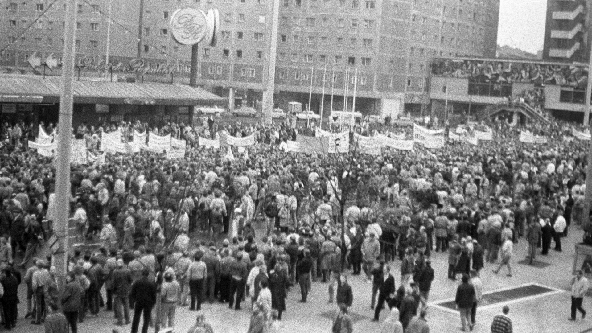Foto, aufgenommen von einer erhöhten Position, von einem Demonstrationszug. Die Demonstranten tragen mehrere Transparente mit nicht zu erkennenden Aufschriften mit sich. Am Rand der Demonstration hat sich eine Gasse von Schaulustigen gebildet.