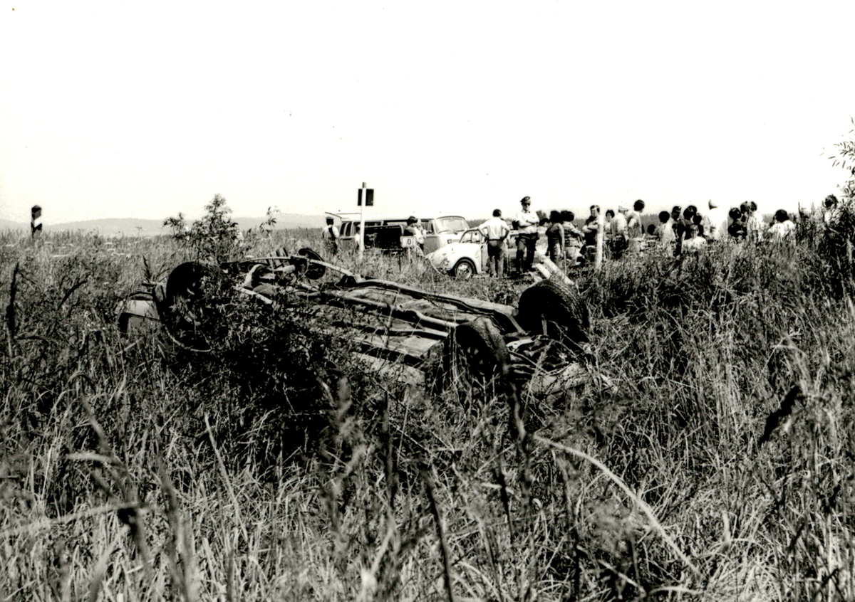 Das auf dem Dach liegende Unfallauto befindet sich abseits der Straße auf einer Wiese. Im Hintergrund befindet sich in einiger Entfernung vom Unfallwagen eine Gruppe Menschen. Am linken oberen Bildrand ist eine DDR-Grenzsäule zu sehen, die die Staatsgrenze der DDR markiert.