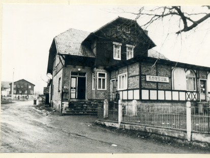 BStU, MfS, HA VII, Nr. 1626, Bl. 12  [Es handelt sich um ein aufgeklebtes schwarzweiß Foto. Den Hauptteil nimmt ein mehrstöckiges Fachwerkhaus in einer langgezogenen Kurve einer unbefestigten Straße ein. An dem flachen Anbau zeigt ein großes Schild den Schriftzug "Konsum". Davor scheint die Einfahrt zum eingezäunten Gelände zu sein. Im Hintergrund sind zwei weitere Häuser zu sehen.]  [Stempel: 3]  Übersichtsaufnahme von der Konsumgaststätte in Bockstadt, in der am 16.12.1975 eine unbekannte Person 2 Bockwürste kaufte.