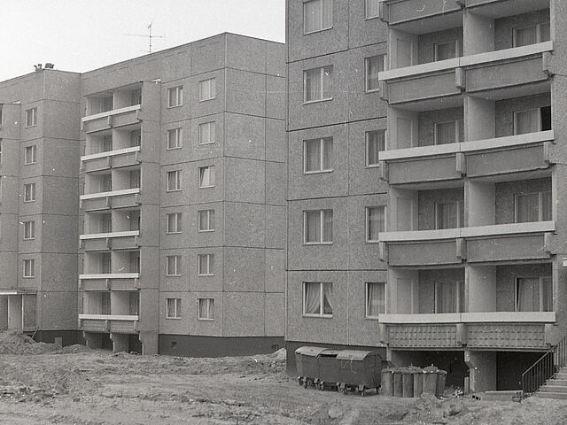 [Die schwarzweiße Negativaufnahme zeigt anteilig zwei sechsstöckige Plattenbauten, vermtulich vom Wohnbauserientyp 70. Sie scheinen bereits bewohnt zu sein, da in allen sichtbaren Fenstern Gardinen hängen. Vor den Wohnhäusern stehen mehrere Mülltonnen aus Metall. Die angrenzenden Freiflächen vor und zwischen den beiden Häuserblocks wirken baustellenartig, Furchen ziehen sich durch das Sand-Geröll-Gemisch. In der unteren rechten Bildecke ist eine große liegende Kabeltrommel.]
