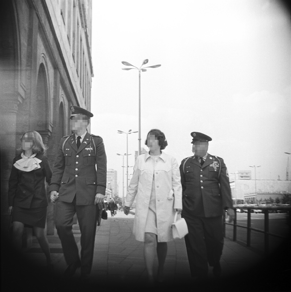 [Zu sehen ist ein schwarz-weißes Negativ, das im Mittelpunkt zwei Mitglieder der US-Armee in Uniform mit ihren weiblichen Begleitungen zeigt. Sie gehen dem Fotografierenden zugewandt über einen breiten Gehweg. Das Bild ist an den Ecken verdunkelt, was den Eindruck erweckt, dass die Kameralinse bei der Aufnahme hinter einem runden Ausschnitt verborgen war.]
