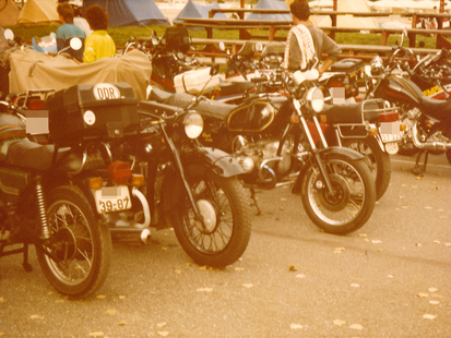 Zu sehen ist ein farbiges Lichtbild. Auf einer asphaltierten Straße sind viele Motorräder dicht nebeneinander abgestellt sind. Einige sind mit einem DDR-Schild gekennzeichnet. Vereinzelt sind Personen dazwischen. Im Hintergrund ist eine grüne Wiese mit aufgestellten Campingzelten zu sehen.