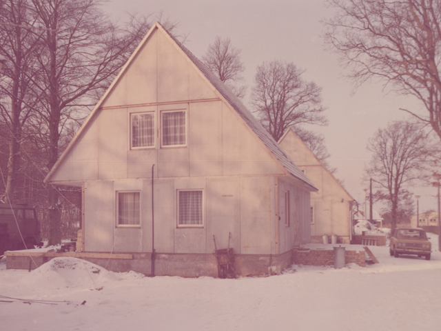 [Das Foto zeigt ein Grundstück mit zwei noch nicht fertiggestellten, baugleichen zweigeschossigen Häusern mit Spitzdächern. Die Dächer, der Boden des Areals und umliegende Sandhügel sind mit Schnee bedeckt. Die Fassaden der beiden Häuser sind noch unverputzt. Neben der Terrasse des vorderen Hauses steht eine Mülltonne aus Zink. Zwischen beiden Häusern auf dem parkt ein gelber Lada.]