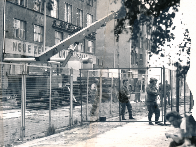 Das schwarz-weiße Lichtbild zeigt das Gebäude der "Neuen Zeit" vom Union Verlag, davor steht ein Schwerlastkran. Hinter der Abgrenzung durch einen Bauzaun stehen mehrere Männer, teils uniformiert und teils mit Bauhelmen. Das Foto war mittig zerrissen worden und ist nun manuell rekonstruiert, Auf der rechten Seite fehlt ein Stück.