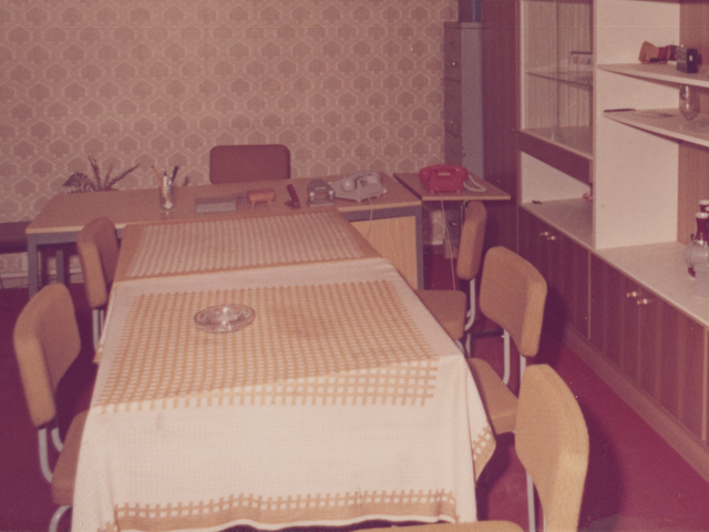 [Die Farbaufnahme zeigt einen Besprechungsraum. Er ist mit rotem Teppichboden ausgelegt. An der linken Raumseite steht fast über die gesamte Länge eine braune Schrankwand aus Holz mit zahlreichen Türen und einer Glasvitrine. Hinter der Schrankwand steht in der Ecke ein Aktenschrank aus Metall. Auf ihm steht ein schwarzer Aktenkoffer. Parallel zur hinteren Wand steht ein Schreibtisch, dahinter ein Stuhl. Auf der Schreibtischoberfläche befinden sich neben einem roten und einem grauen Schnurtelefon mehrere Büro-Utensilien. Vor dem Schreibtisch stehen zwei quadratische Tische, die mit Tischdecken behangen sind. Rund um die Tische stehen fünf Stühle. mAuf dem vorderen Tisch steht ein Aschenbecher. Über den Tischen hängt eine Lampe mit fünf bauchig geformten Lampenschirmen aus Glas.]