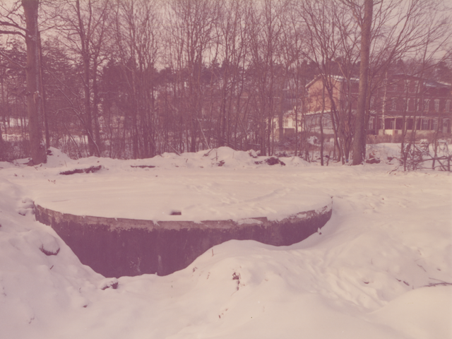 [Im Vordergrund der Farbaufnahme ist ein großer runder Block aus Stein zu sehen, unter dem sich vermutlich eine Klärgrube verbirgt. Der gesamte Deckel sowie die umliegenden Bodenflächen sind mit Schnee bedeckt. Durch kleine, unbeblätterte Bäume hindurch sind mehrere Gebäude erkennbar. Im Bildhintergrund rechts ist ein breites dreigeschossiges Haus mit einem flachen Spitzdach und einer überdachten Terrasse zu sehen.]