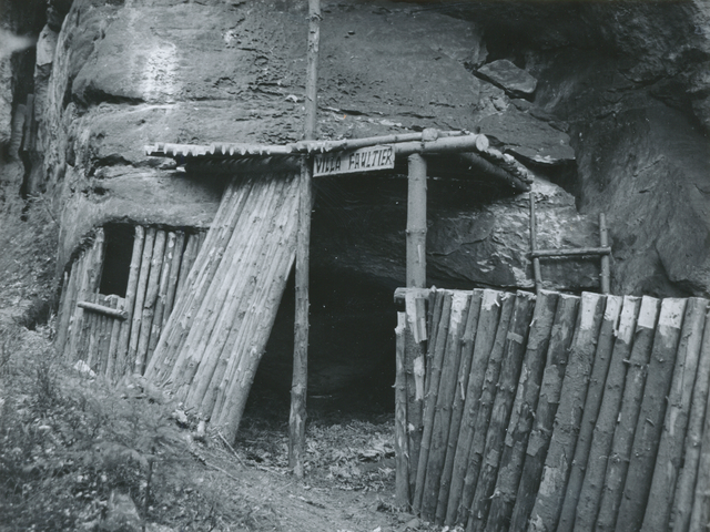 Die Schwarzweißfotografie bildet das Äußere der Boofe ab. Man sieht dort den hüfthohen, begrenzenden Zaun aus dünnen Baumstämmen und den überdachten Eingang, an dem ein Schild mit der Aufschrift "VILLA FAULTIER" angebracht ist. 