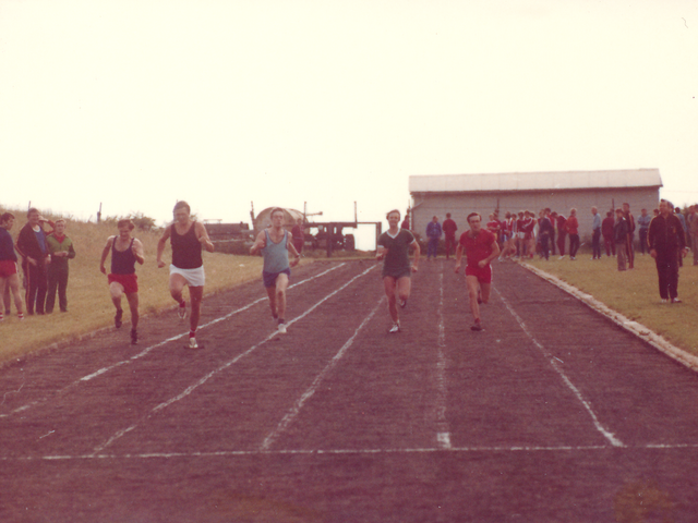 [Fünf Sprinter sind auf der Laufbahn zu sehen. Im Hintergrund und am linken Rand schauen ihnen weitere Sportler zu. Im Hintergrund ist eine weiße eingezäunte Baracke zu sehen.]