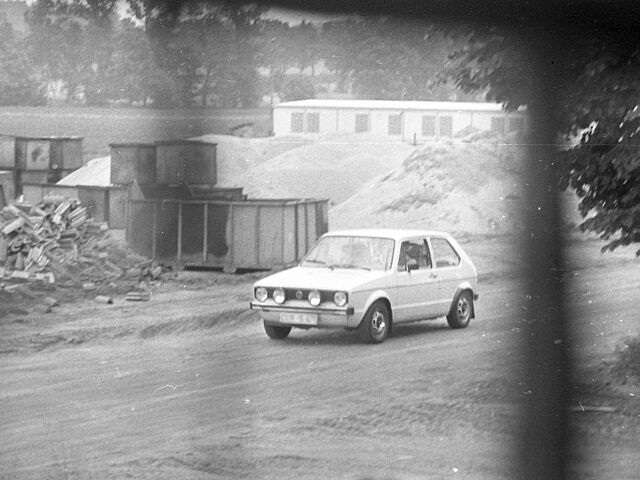 [Das schwarzweiße Lichtbild zeigt einen hellen VW Golf I, wie er auf einem Sandweg vermutlich auf einer Baustelle entlang fährt. Im Hintergrund ist links eine Anhäufung von Dachziegeln zu sehen, daneben wurden mehrere leere Container übereinander gestapelt. Dahinter befinden sich mehrere Sandberge vor einem Flachbau.]