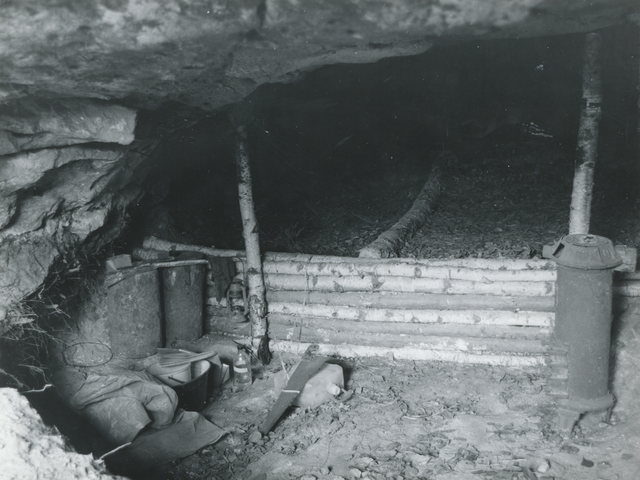 Das Bild, eine Schwarzweißfotografie, zeigt das Innere der Boofe. Im Vordergrund sind auf der linken Seite Töpfe und anderes Kochgeschirr zu sehen und rechts ein weiteres längliches Gerät aus Metall. Hinter einer niedrigen Absperrung aus Birkenästen geht es tiefer in den Fels hinein, wo der Boden mit Laub ausgelegt ist.