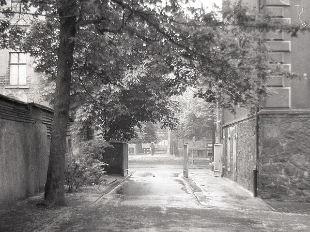 [Vermutlich handelt es sich um einen Innenhof, der zu einem befestigten Parkplatz ausgebaut wurde. Zu sehen ist die betonierte Einfahrt durch ein offenstehendes Tor, rechts daneben steht das vermeidliche Tor für Fußgänger ebenfalls offen. Der Parkplatz liegt hinter einem Altbau, dessen Hausecke auf der rechten Seite angeschnitten wird, während auf der linken Seite eine mannshohe Gartenmauer verläuft. Der Blick vom Gelände hinunter zeigt einen breiten Gehweg auf der gegenüberliegenden Straßenecke. Es ist ein schwarzweißes Negativ, dass im rechten unteren Drittel durch einen Riss beschädigt ist.]