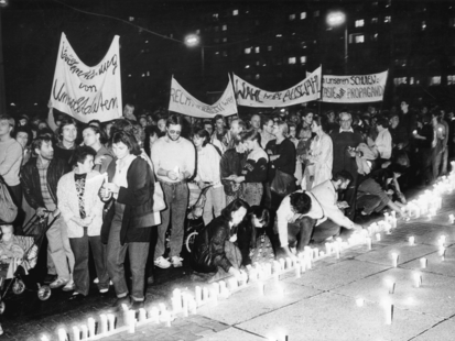 [Eine Reihe brennender Kerzen wird an einem Treppenabsatz aufgestellt, einige Menschen sind dabei  auf schwarzweißem Foto abgelichtet worden. Dahinter ziehen Demonstranten mit Transparenten vorbei.]