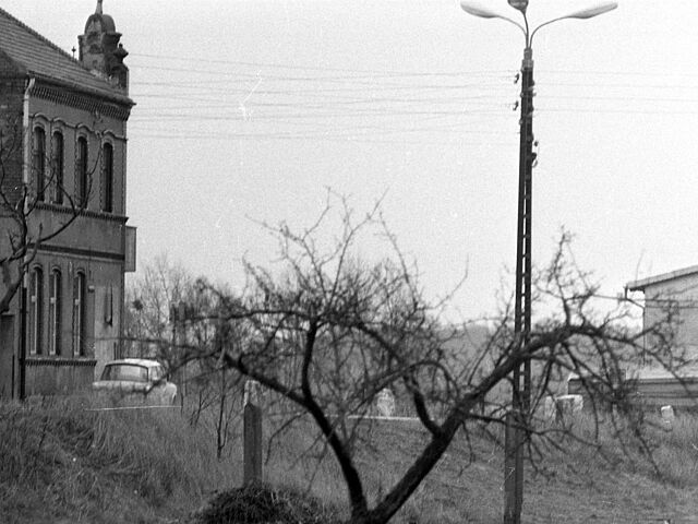 [Das Schwarzweiß-Positiv zeigt die Ecke eines merkstöckigen Backsteinhauses. Davor ist ein heller Trabant zu sehen. Markant ist rechts davon ein Strommast, der gleichzeitig als dreistrahlige Straßenlaterne dient. Im hintergrund ist ein Teil der Stirnseite eines langen Flachbaus zu sehen.]