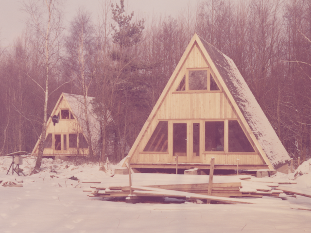 [Das Farbfoto zeigt zwei Finnhütten vor einem Waldstück. Boden und Dächer sind mit Schnee bedeckt. Die Dachflächen reichen bis zum Boden, die Frontflächen sind holzgetäfelt und mit Türen und Fenstern, von denen ein dreigeteiltes Fenster zur oberen Etage gehört, versehen. Vor der vorderen Hütte liegen aufgestapelte Holzleisten.]