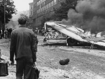 Der Betrachter des Schwarz-Weiß-Bildes sieht einen brennenden Bus. Das Dach des Busses ist in sich zusammengefallen. Teile der Verwüstung liegen auf der Straße. Vorne links im Bild stehen zwei Männer mit Aktentaschen, die das Geschehen beobachten. Im Hintergrund, entlang der Straße sind Menschen zu erkennen.