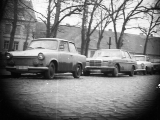 In Nienburg (Saale) wurden mit versteckter Kamera am Straßenrand parkende Trabants und ein Mercedes aufgenommen (mit Blick auf die Gattersleber Straße). Im Hintergrund sind Häuser und Bäume zu sehen.