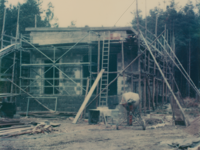 [Das Farbfoto zeigt den vollständig eingerüsteten Rohbau eines eingeschossigen Hauses. Vor dem Gebäude steht ein Betonmischer, es liegen verschiedene Baumaterialien wie Planen, Holzleisten und Steine herum. Am Gerüst lehnt eine Leiter, die bis zum flachen Schrägdach reicht. Links ist ein Förderband aufgestellt. Hinter dem Gebäude stehen Nadelbäume.]
