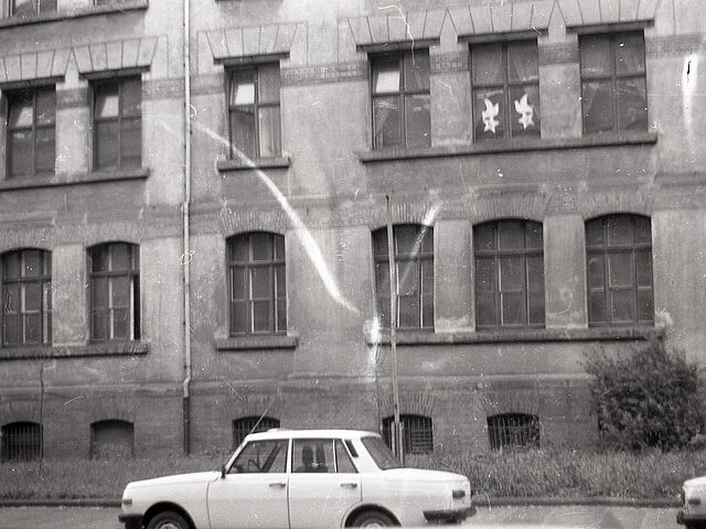 [Das schwarzweiße Negativ zeigt eine unscharfe Aufnahme von den ersten zwei Stockwerken eines Mietshauses. Vor dem Haus verläuft ein schmaler Grünstreifen, gefolgt von einem befestigten Gehweg. Es parken zwei helle Lada am Straßenrand, vom rechts gelegenen ist nur der Frontteil zu sehen. In der rechten oberen Hälfte verläuft ein kleiner Riss durch das Fotopapier, eine Spiegelung in der Mitte des Bildes lässt auf weitere Deformierungen des Bildes schlussfolgern.]