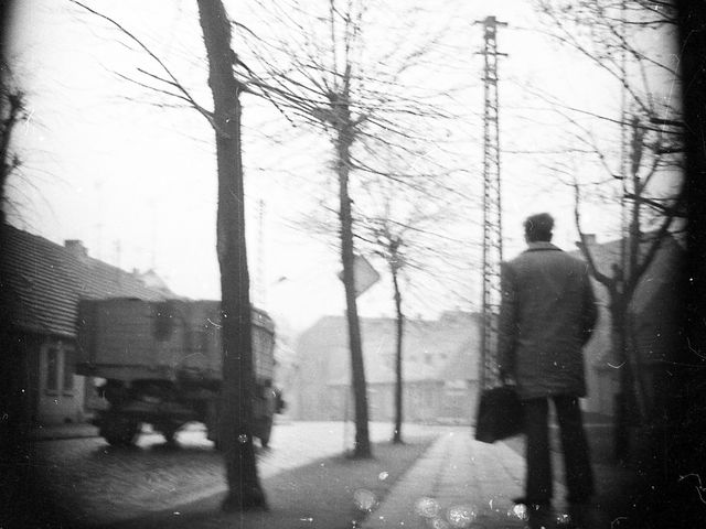 Die Aufnahme mit versteckter Kamera zeigt die Gattersleber Straße in Nienburg (Saale), auf der ein LKW entlangfährt. Auf dem Gehweg ist die Rückseite eines Mannes mit oberschenkellanger Jacke und Aktentasche in der linken Hand zu sehen. Rechts und links der Straße stehen Einfamilienhäuser.