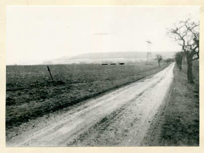 BStU, MfS, HA VII, Nr. 1626, Bl. 11   [Ein aufgeklebtes schwarzweißes Fotopositiv, das im Zentrum eine unbefestigte Straße zeigt. An den Straßenrändern stehen vereinzelte kahle Bäume. Links davon ein brach liegendes Gelände. Ein eingezeichneter Pfeil deutet auf ein winziges Gebäude an der Horizontlinie, auf der auch ein gestrichelter Pfeil nach rechts deutend abgebildet ist. Im Bildhintergrund und wie im Nebel wirkend sind bergige Landschaftserhebungen zu sehen.]  [Stempel: 2] Auf dem Foto ist die Straße von Bürden nach Veilsdorf abgebildet. Auf dieser Straße im PKW fahrend sahen die Zeugen [anonymisiert] und [anonymisiert] eine männliche Person auf dem mit der gestrichelten Linie markierten Geländeabschnitt gehen. Der Pfeil zeigt auf die Milchviehanlage Schackendorf, wo am 15.12.1975 in den Abendstunden der PKW "Wartburg" [anonymisiert] von Weinhold abgestellt wurde.