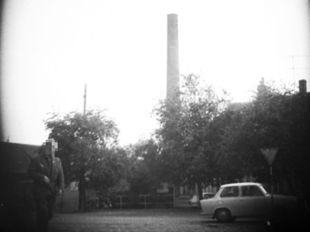 Mit versteckter Kamera wurden ein Mann und ein Trabant auf einem Parkplatz aufgenommen. Hinter Bäumen ragen ein Industrieschornstein, ein Mast und ein Dach hervor.