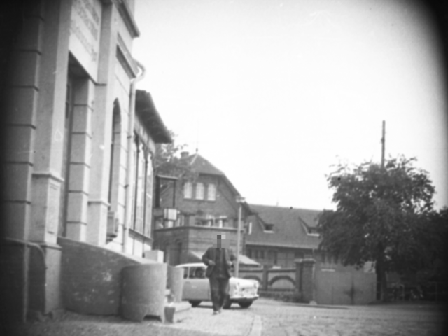 Zu sehen sind Gebäude am Rand einer unbekannten Straße. Ein männlicher Fußgänger passiert eines der Häuser. Aus der Linkskurve im Hintergrund kommt ein Trabant gefahren.