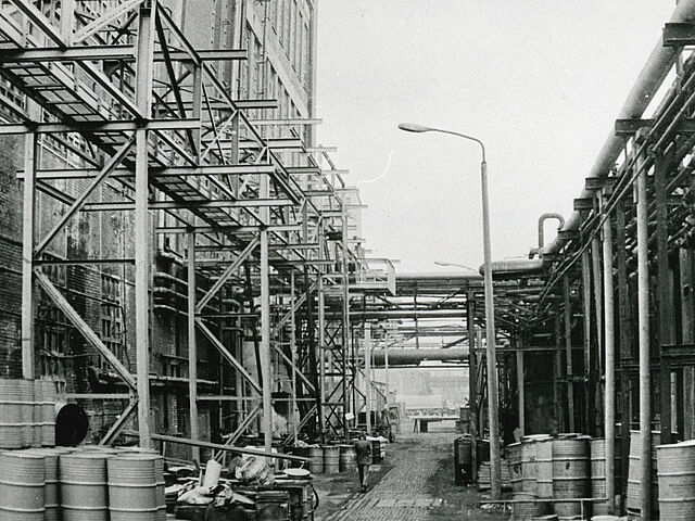 [Auf der schwarz-weiß Fotografie ist ein Teil eines Industriegebietes zu erkennen. Links ist ein mehrstöckiges Gebäude zu sehen, welches von einem Stahlgerüst umgeben ist. In der Mitte ist ein gepflasterter Weg zu erkennen, auf dessen linker Seite eine Person entlanggeht. Rechts am Weg entlang stehen Straßenlaternen. Auch ist auf dem Boden eine Mehrzahl von Tonnen zu sehen. Rechts neben dem Weg steht vermutlich eine Mauer, an der einige Rohre entlang führen. Im Hintergrund sind weitere Industriegebäude zu erkennen.]