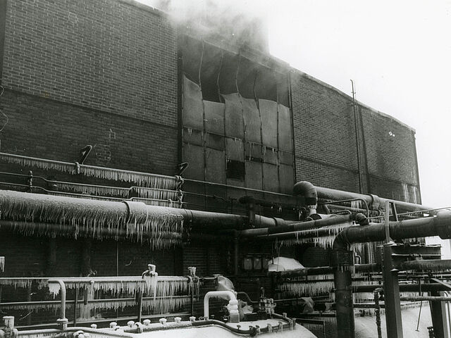[Auf der schwarz-weiß Fotografie ist ein Teil des Gebäudes von Bild 17 und Bild 20 zu sehen. In der Mitte des Backsteingebäudes gibt es einen Abschnitt, der aus Metallplatten besteht. Einige der Platten sind stark deformiert und lassen Dampf aus dem Inneren des Gebäudes entweichen. Vor dem Gebäude verlaufen einige Rohre. Am Fuß des Gebäudes sind zwei waagerechte Tanks zu erkennen. Durch die starke Eiszapfenbildung an den Rohren und des Schnees auf den Tanks ist es wahrscheinlich, dass das Foto im Winter aufgenommen wurde.]